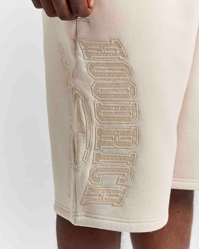 Close-up of a person wearing cream-colored athletic shorts with large tonal embroidered lettering on the side.