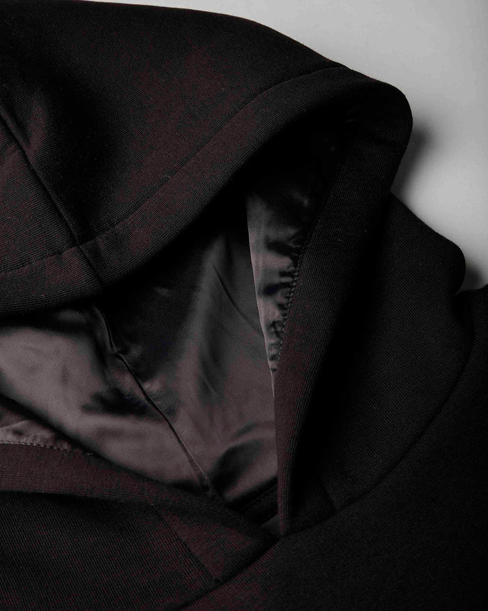 Close-up detail of a black hooded jacket or coat with a satin-like black interior lining, shown against a light gray background.