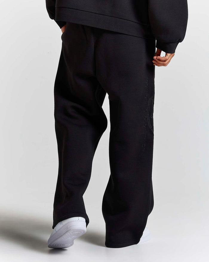 A person shown from the back wearing loose black sweatpants and a black sweatshirt, with white sneakers, against a light gray studio background.