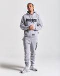 A man modeling a light gray Hoodrich hoodie and matching sweatpants set, featuring a large black and blue 'HOODRICH' logo across the chest and smaller branding on the pants, against a light gray studio background.