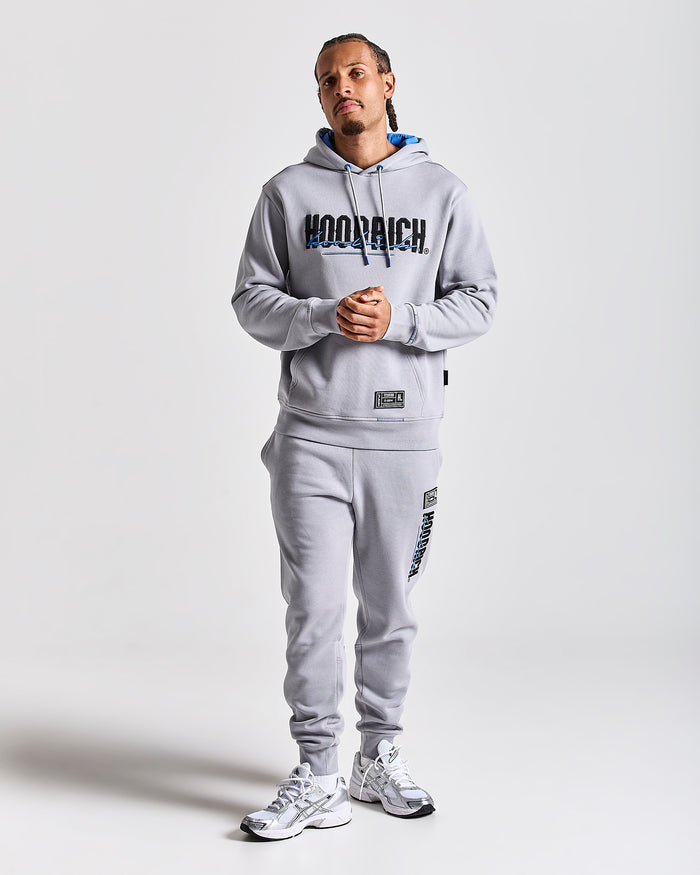 A man modeling a light gray Hoodrich hoodie and matching sweatpants set, featuring a large black and blue 'HOODRICH' logo across the chest and smaller branding on the pants, against a light gray studio background.