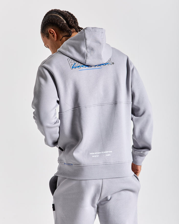 A man wearing a light gray pullover hoodie and matching sweatpants, shown from the back against a light gray studio background. The hoodie features a large blue and white graphic on the upper back and small white text near the lower back.