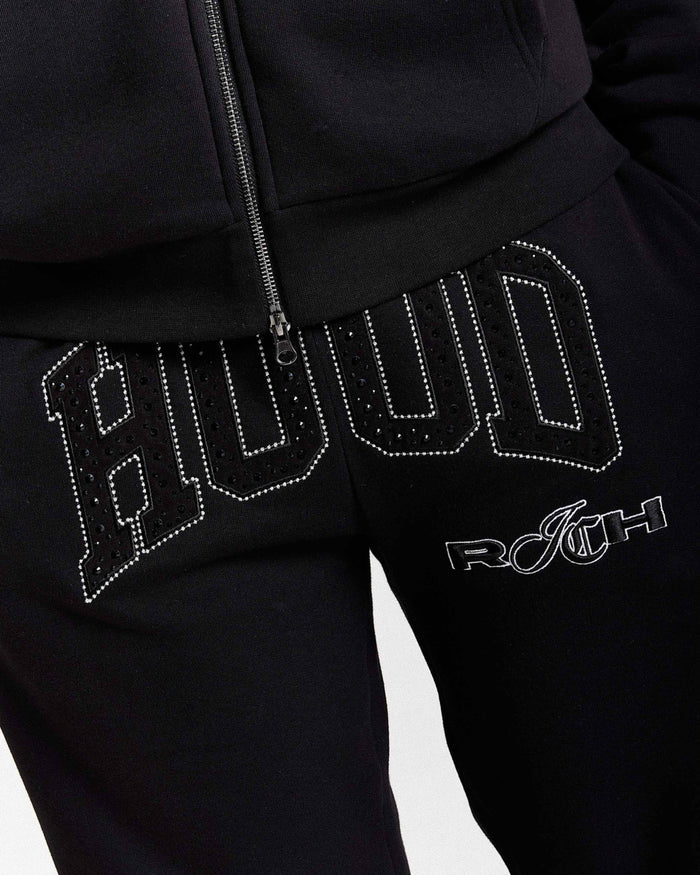Close-up of black sweatpants with large rhinestone-studded 'Hood' lettering across the thigh and a white 'Rich' logo below, paired with a black zip-up top.