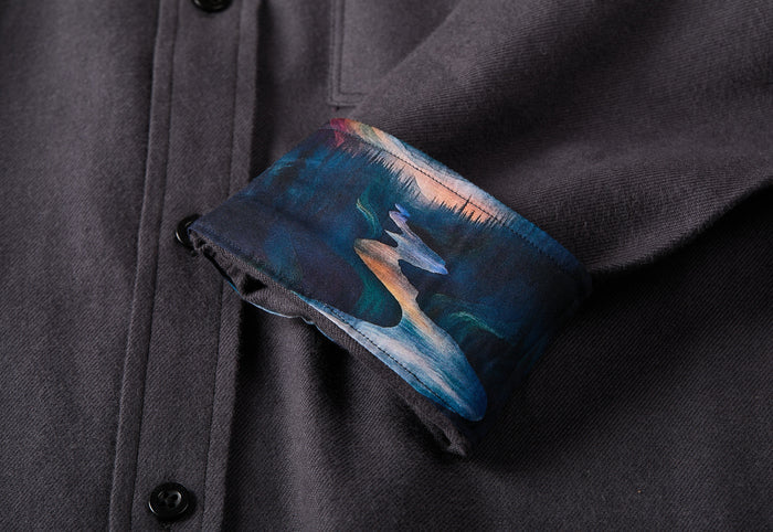 The Men's Cascade Flannel Shirt in Charcoal Grey is a modern-cut, Portuguese cotton shirt with cuffs lined inside with a vibrant landscape art depicting a river, trees, and sunset.