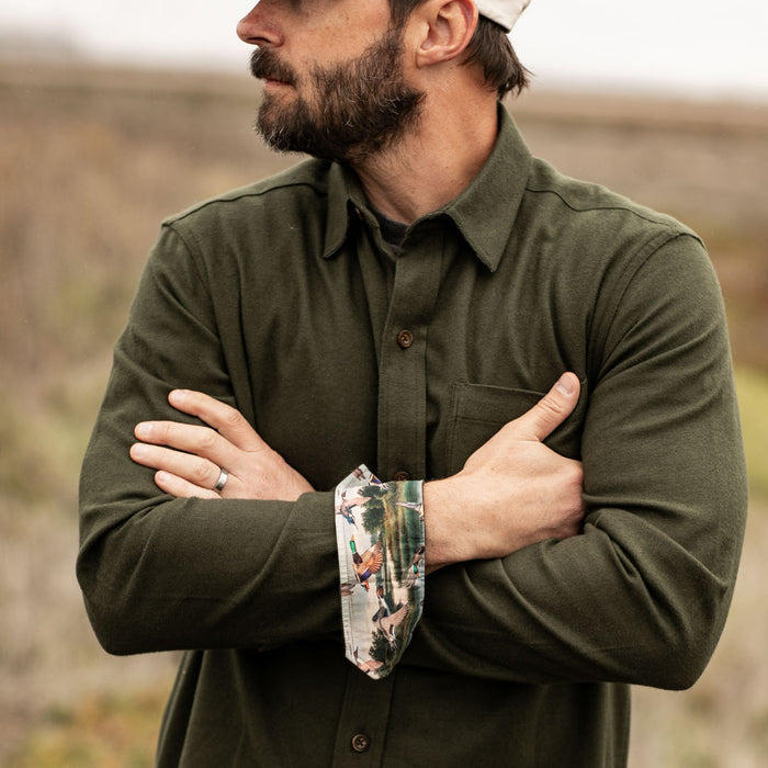 Bearded man outdoors, arms crossed, wearing Pladra Men's Cascade Flannel Shirt in Olive Green and a cap.