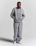 A man modeling a light gray two-piece tracksuit consisting of a lightweight zip-up jacket and matching jogger pants, paired with white sneakers, against a plain light gray studio background.