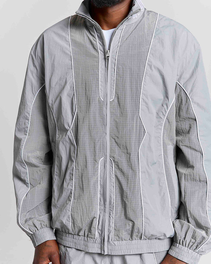 A close-up of a person wearing a light gray zip-up windbreaker jacket with white piping and a subtle grid texture, paired with matching gray pants against a plain light background.