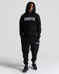 A man modeling a black hoodie and sweatpants set with white 'HOODRICH' branding, standing against a light gray studio background.