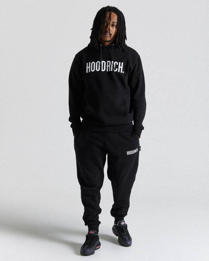 A man modeling a black hoodie and sweatpants set with white 'HOODRICH' branding, standing against a light gray studio background.