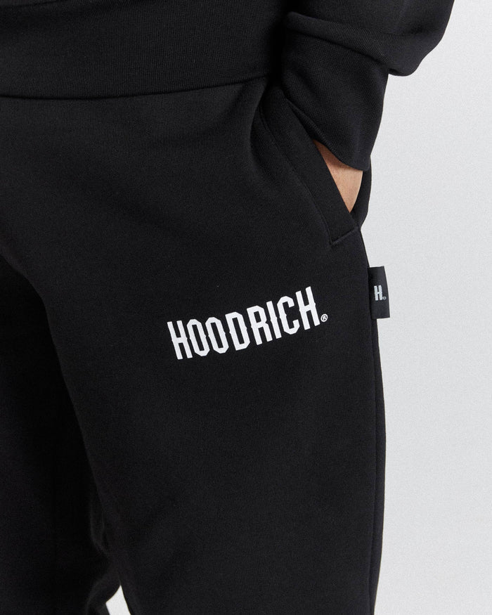 Close-up of black sweatpants with a white 'HOODRICH.' logo printed on the thigh and a small black side tag, shown on a model against a light gray background.