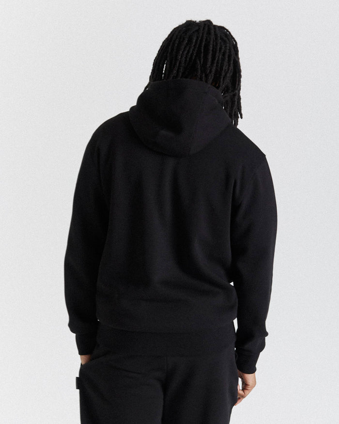 A person viewed from the back wearing a black pullover hoodie and black pants against a plain light gray studio background.