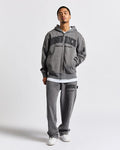 A man modeling a gray zip-up hoodie and matching sweatpants set from Good Rich, featuring black collegiate-style lettering on the front and thigh, styled with white sneakers against a light gray studio background.