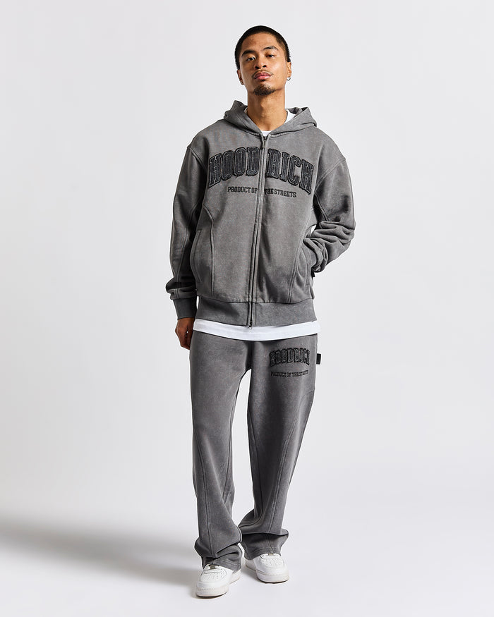 A man modeling a gray zip-up hoodie and matching sweatpants set from Good Rich, featuring black collegiate-style lettering on the front and thigh, styled with white sneakers against a light gray studio background.