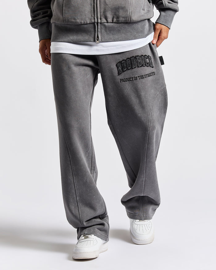 A person wearing gray washed sweatpants with black 'HOODRICH' and text on the left thigh, styled with a white undershirt and white sneakers against a light gray studio background.