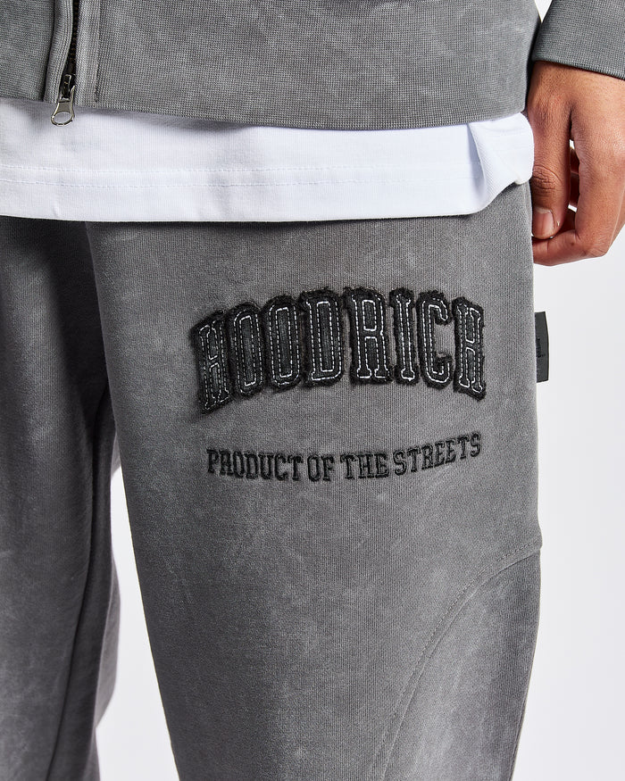 Close-up of a person wearing gray hooded sweatpants with black embroidered text reading 'HOODRICH' on the thigh, plus a white undershirt and gray zip jacket visible.
