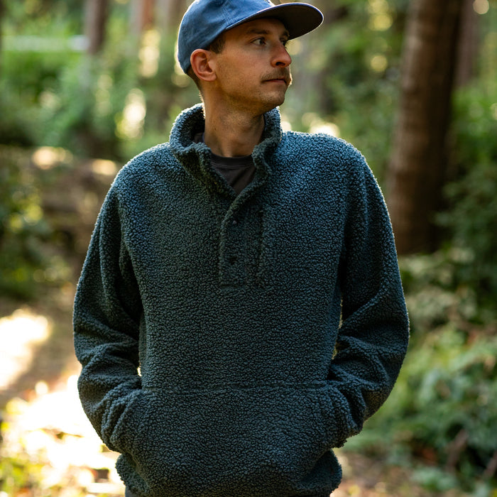 Man wears Pladra Men's Cypress Sherpa Fleece in Atlantic Blue and cap, standing outdoors in a sunlit forest.