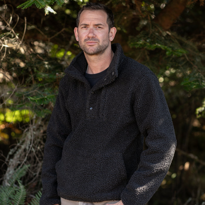 A man with short dark hair and a trimmed beard stands outdoors in front of sunlit green trees, wearing the Men's Cypress Sherpa Fleece in Onyx Black.