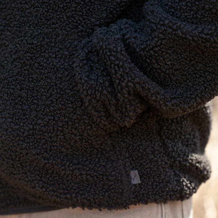 A close-up shows a person wearing the Men's Cypress Sherpa Fleece in Onyx Black, featuring soft curly texture and a small rectangular tag near the hem, with their hand tucked into the pocket.