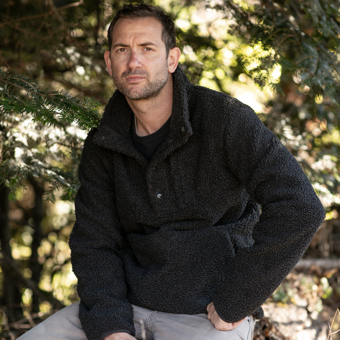 A man with short dark hair and stubble sits outdoors among green trees, wearing the Men's Cypress Sherpa Fleece in Onyx Black and light pants, looking directly at the camera with a neutral expression.