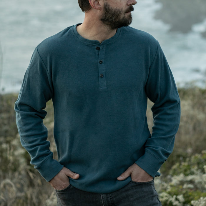 A bearded man stands outdoors with his hands in his pockets, wearing the Men’s Elko Henley in Atlantic Blue and dark jeans, with blurred greenery and water in the background.