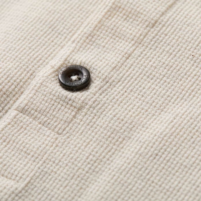 Close-up of the Men's Elko Henley in Oatmeal White, showing its textured cream waffle fabric and a black button bearing engraved text around the edge.