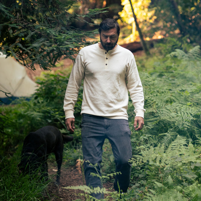 A man in the Men's Elko Henley - Oatmeal White and jeans walks along a fern-lined forest path with a dark-colored dog, as sunlight filters through the trees.