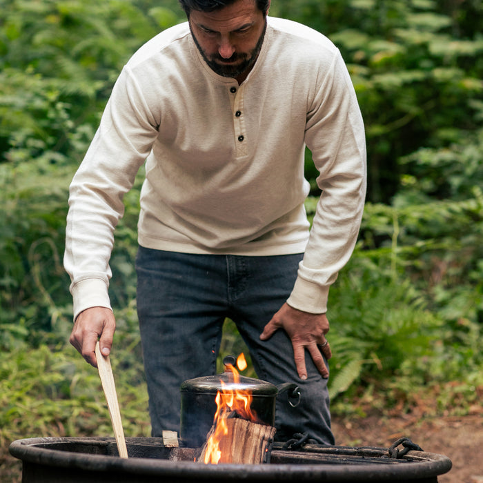 A man wears the Men's Elko Henley in Oatmeal White as he stirs a pot over an open campfire, surrounded by trees and greenery.