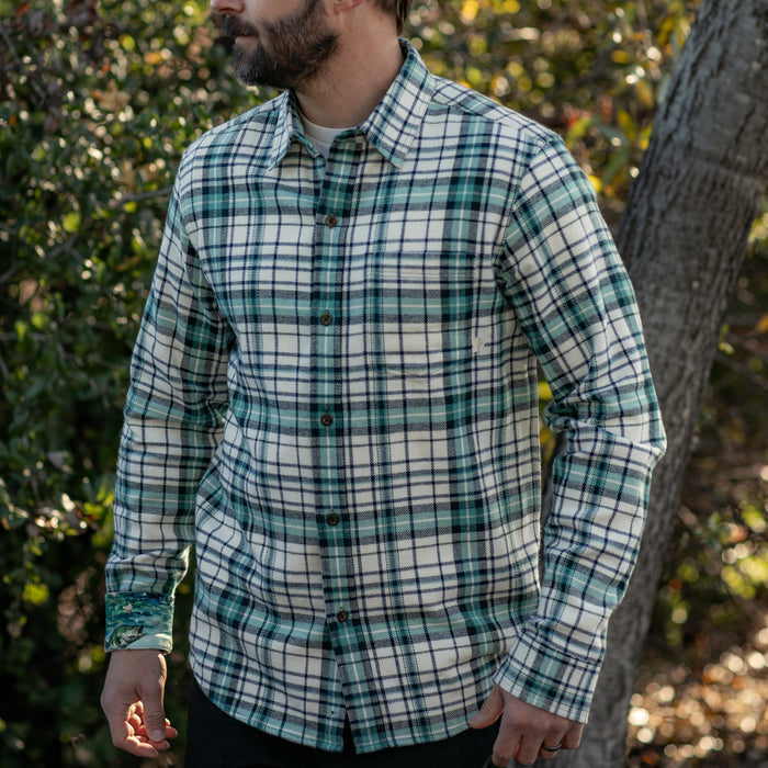 A man wears a Pladra Men's Every Day Elite Flannel Shirt in Everglade White, standing outdoors by trees and bushes.