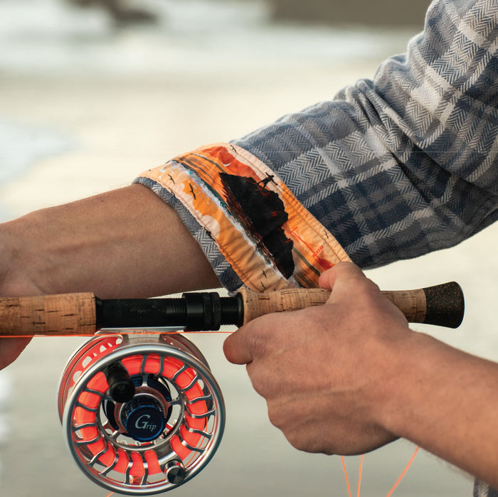 A person wearing the Men's Every Day Elite Flannel Shirt in Geary Grey holds a fly fishing rod with both hands. The heavyweight cotton flannel cuff and the orange and silver fishing reel stand out.