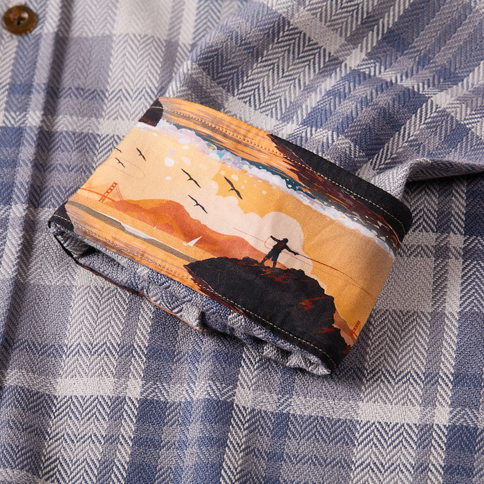 The Men's Every Day Elite Flannel Shirt- Geary Grey is a blue and gray plaid heavyweight cotton flannel, featuring turned-up cuffs that reveal a unique inner lining with illustrations of hills, a bridge, birds, and a person with arms raised.