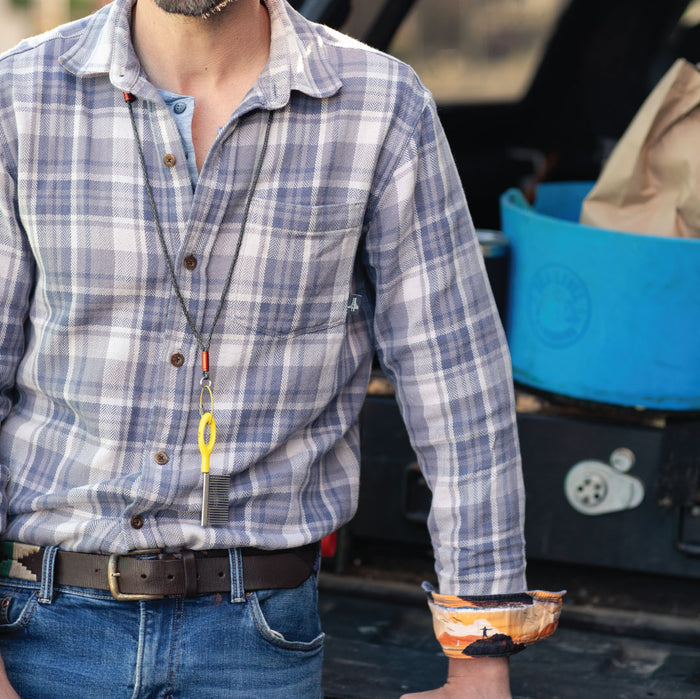 A man in the Men's Every Day Elite Flannel Shirt- Geary Grey and jeans stands by a vehicle with a blue container and paper bag in back. The heavyweight cotton shirt has rolled-up sleeves with colorful lining, and he wears a necklace with a yellow tool.