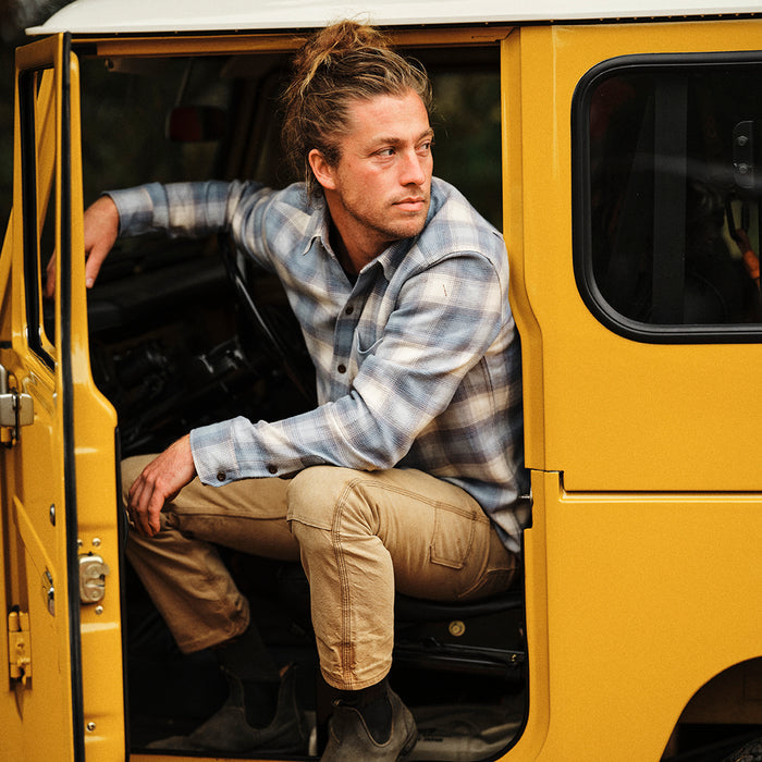 A man with long hair tied back, wearing the Men's Every Day Elite Flannel Shirt in Mendocino Blue and khaki pants, sits sideways in a yellow vehicle's open doorway, gazing thoughtfully into the distance.