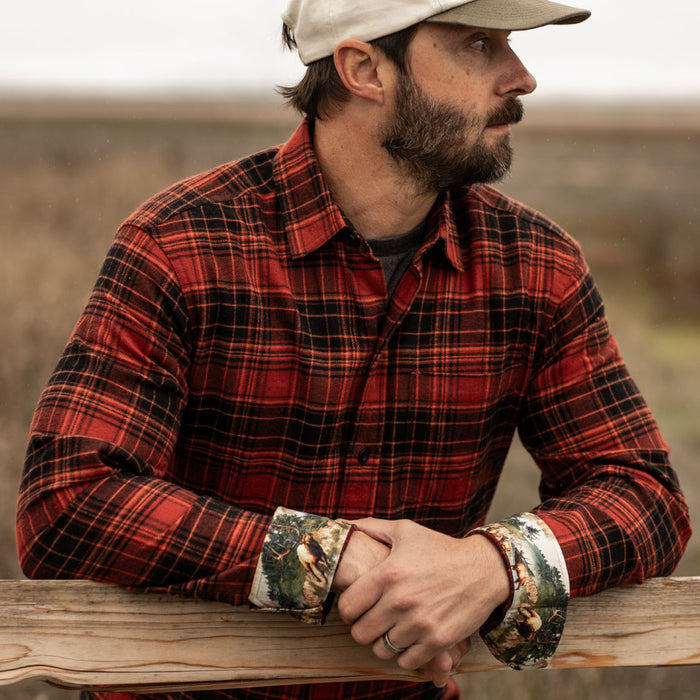A man in the Men's Every Day Flannel Shirt- Autumn Red and a beige cap leans on a wooden fence, gazing into the distance in an outdoor rural setting.
