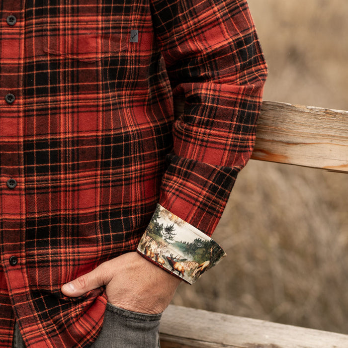 A person stands by a wooden fence with one hand in their pocket, wearing the Men's Every Day Flannel Shirt- Autumn Red. The shirt cuff is turned up to reveal a scenic nature illustration on the inside.
