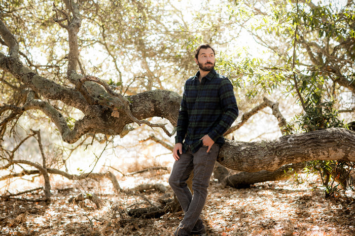 A bearded man in the Men's Every Day Flannel Shirt- Bolinas Blue and jeans sits on a large tree branch in a sunlit forest, surrounded by leaves and dappled sunlight.