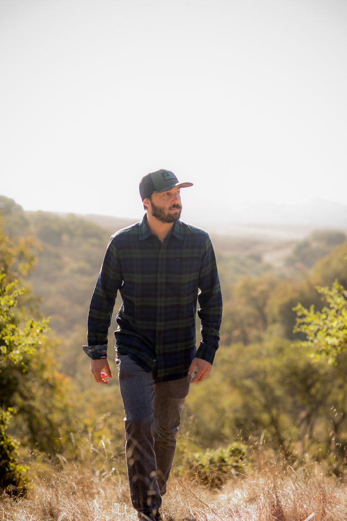 A bearded man in a Men's Every Day Flannel Shirt–Bolinas Blue and a baseball cap walks through dry grass on a sunny day, hills in the background.