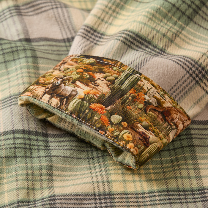 A close-up of the Men's Every Day Flannel Shirt- Cactus Green shows its folded green plaid design and a fabric cuff featuring a colorful desert scene with cacti, flowers, deer, and birds—perfect for everyday wear with unique style.