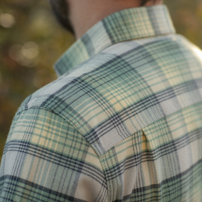 Close-up of a person wearing the Men's Every Day Flannel Shirt in Cactus Green, highlighting its green, white, and blue pattern and soft fabric texture, with a blurred outdoor background. The head and neck are partially visible.