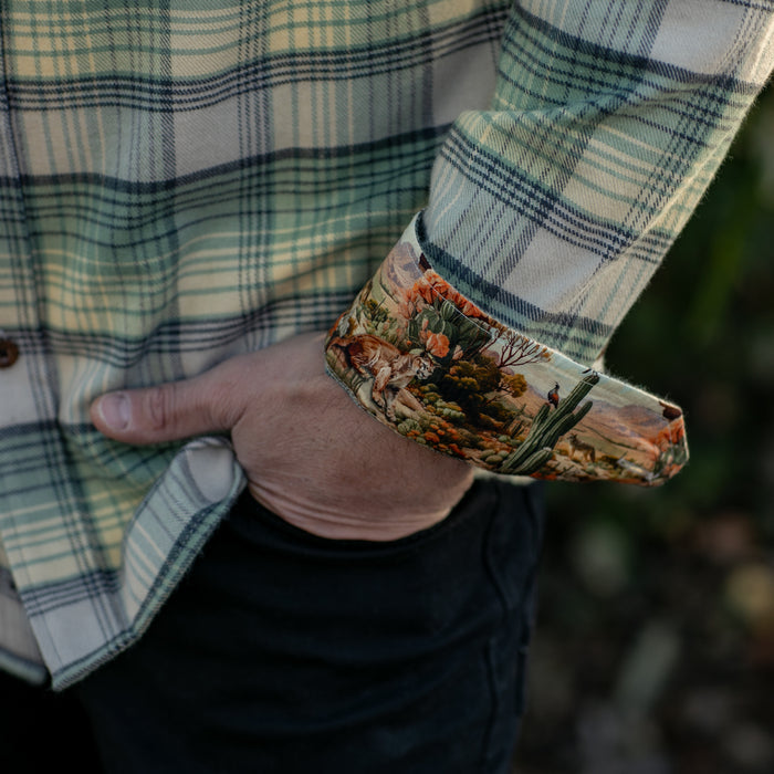 A person wearing the Men's Every Day Flannel Shirt- Cactus Green shows off a vibrant desert landscape pattern on the inner cuff with rolled sleeves, pairing it effortlessly with black pants and one hand tucked in a pocket.