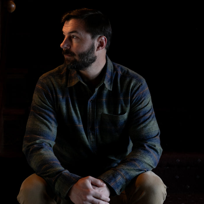 A bearded man with short dark hair, wearing the Men's Every Day Flannel Shirt in Everett Green Heather and tan pants, sits with his hands clasped, looking to the left against a dark background.