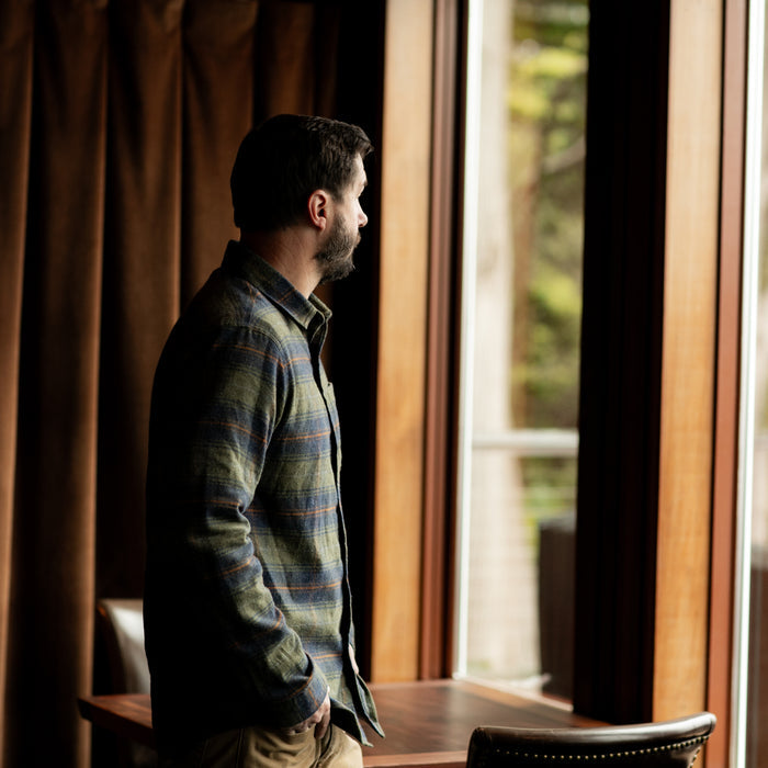 A man with dark hair and a beard stands indoors by a large window, wearing the Men's Every Day Flannel Shirt in Everett Green Heather. Brown curtains, a wooden table, and soft natural light complete the cozy setting.