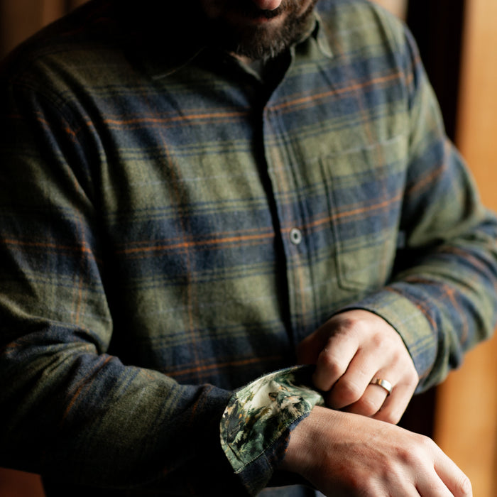 A man adjusts the cuff of the Men's Every Day Flannel Shirt in Everett Green Heather, revealing its camouflage-patterned lining. His wedding ring is visible as he showcases this stylish and rugged outdoor shirt.