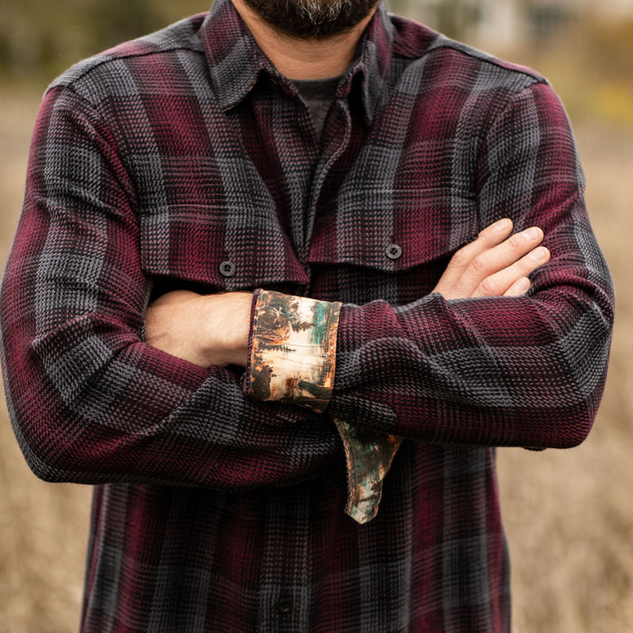A bearded man stands outdoors with his arms crossed, wearing the Men's Fireside Flannel - Hickory Red. The rolled-up cuffs reveal scenic print fabric, and tall grass is visible in the blurred background.