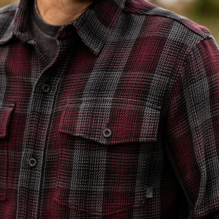 A close-up of a person wearing the Men's Fireside Flannel in Hickory Red—a plaid shirt with burgundy, black, and gray tones over a dark t-shirt. The shirt has two chest pockets with button flaps.