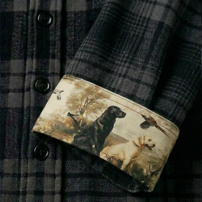 A close-up of the Men's Fireside Flannel - Night Black sleeve, rolled up to show an inner lining with an outdoor scene of a black dog, a yellow dog, flying birds, and trees.