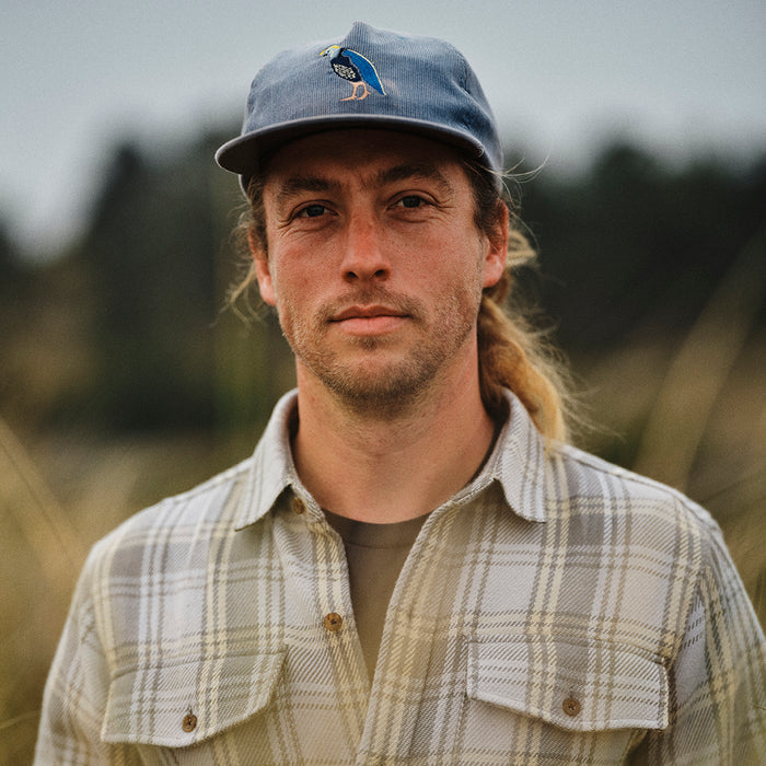 A man with long blond hair in a ponytail, wearing a blue cap and the Men's Fireside Flannel - Quartz Grey, stands outdoors with blurred greenery behind him.