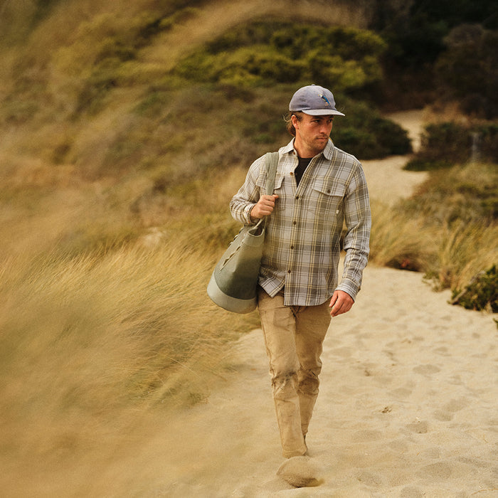 A man in a Men's Fireside Flannel - Quartz Grey, khaki pants, and a cap walks along a sandy trail through grassy dunes, carrying a large green duffel bag over his shoulder.