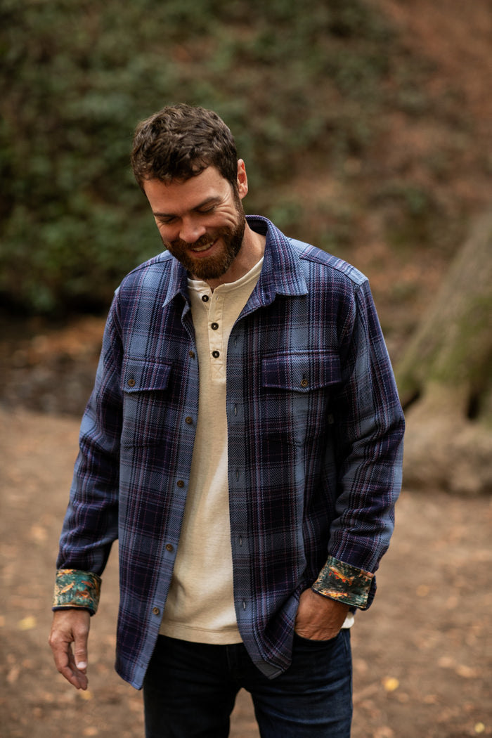 Bearded man in Pladra Men's Fireside Flannel - Riffle Blue smiles, looking down outdoors in the woods.