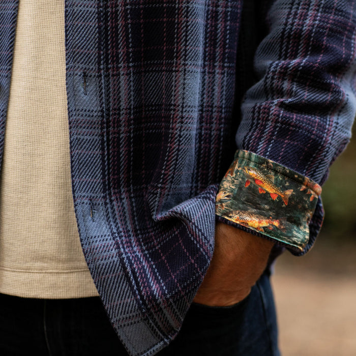 A man in a Pladra Men's Fireside Flannel - Riffle Blue with fish-patterned cuffs stands outdoors, hand in pocket.
