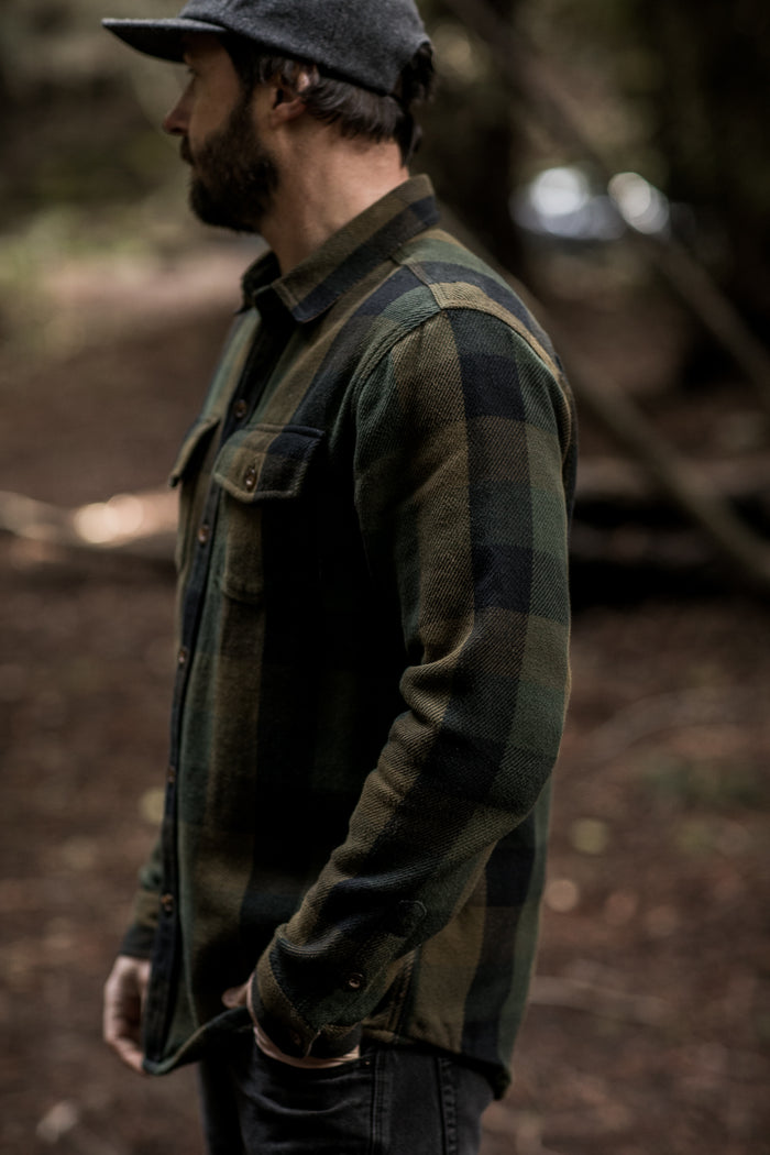Man in a Pladra Men's Fireside Flannel - Woodland Green and gray cap stands sideways in a forest.
